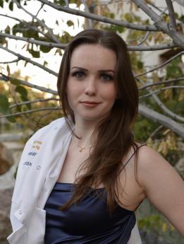 Jillian Cellucci with University of California Davis lab coat over her shoulder, wearing a blue dress and standing by a tree.